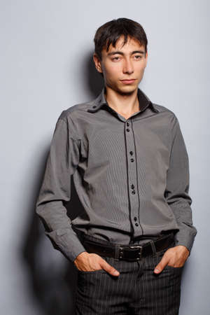 Young man in steel-blue shirt standing on grey background with hands in pocketsの写真素材