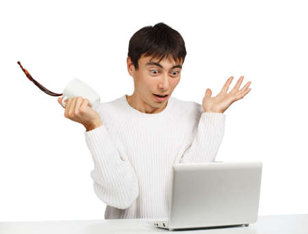 Amazed young man sitting at the netbook with a cup of coffee in his hand, which he spills due to his sudden gesture of surpriseの写真素材