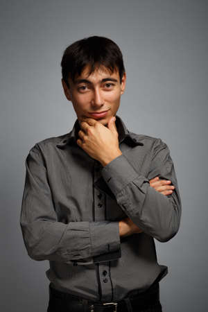 Young man in steel-blue shirt standing on grey background in thoughtful poseの写真素材