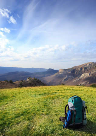 Travel backpack standing on green grass on a background of mountains under the blue cloudy sky