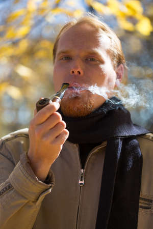 Young beautiful red-haired hipster man smoking tobacco pipe in autumnal park. Shallow DOF.の写真素材