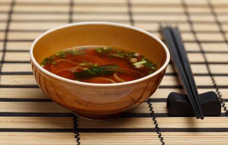 Miso soup in brown bowl standing on bamboo mat with back chopsticks lying near.