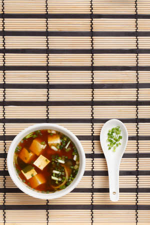 Bowl of delicious traditional japanese miso soup with tofu and wakame seaweed, white ceramic spoon with chopped greens standing on striped bamboo mat.の写真素材