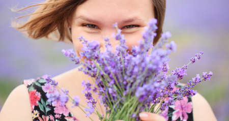 Beautiful young smiling woman with bouquet of lilac lavender flowers dressed in blouse with floral design standing at the background of blurred lavender field.の写真素材