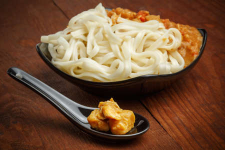 Beautiful hot curry sauce with noodles in black plate standing at brown wooden table with ceramic spoon. Shallow DOF. Focus on spoon.の写真素材