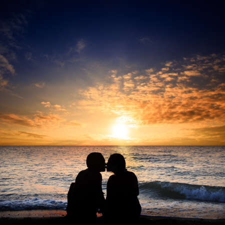 Kissing man and woman sit on the seashore next to the surf zone with small waves under cloudy sky with setting sun at the horizonの写真素材