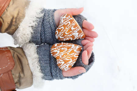 Female hands in knitted grey mittens hold beautiful heart shaped biscuit cookies with white icing at snow background. Focus on cookies. Top view.の写真素材