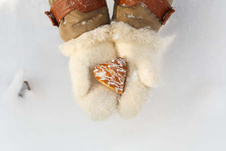 Female hands in woollen white mittens hold beautiful heart shaped biscuit cookie with icing at snow background. Top view.の写真素材