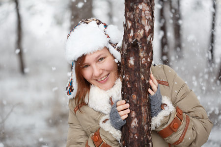 Young beautiful smiling woman in coat with white fur, hat with ear flaps and grey fingerless knitted...