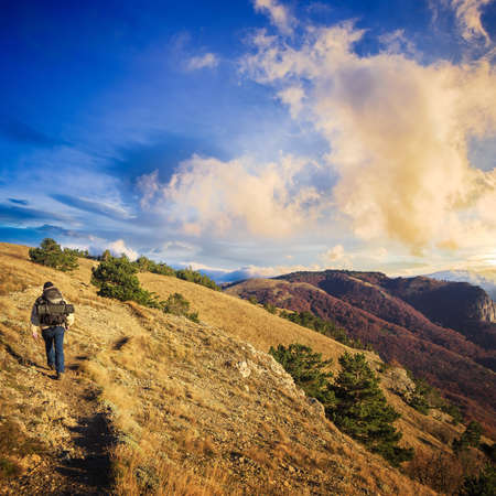 Traveller man with a backpack walks on rocky path at autumnal mountain range with dry grass and sunset sky above.の写真素材