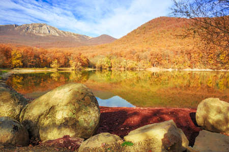 Beautiful scenic mountain lake with autumnal forest and big stones along the shore and blue cloudy sky above and in the reflection in water surface.の写真素材