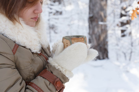Young woman in winter coat and white fluffy mittens holds pottery cup in her hands at the background...