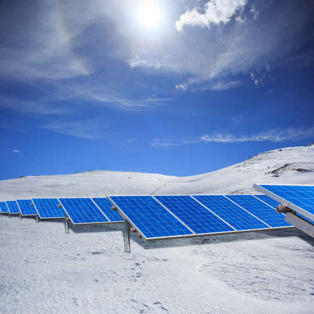 Beautiful modern solar station with blue panels standing in winter field with white snow and hills at the horizon under bright cloudy sky with sunlight.の写真素材