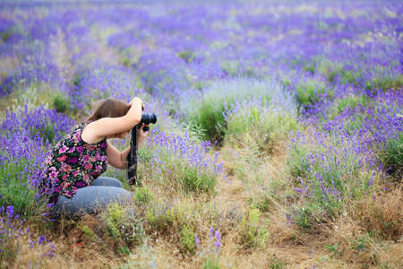 Young beautiful woman in blouse with flower ornament sitting at purple lavender field with photo camera in hands and taking picture.の写真素材