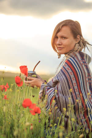 Young beautiful woman in poncho with ethnic pattern sits on field in the middle of green grass and red poppies at the background of cloudy sunset sky and holds calabash with bombilla in hands.の写真素材