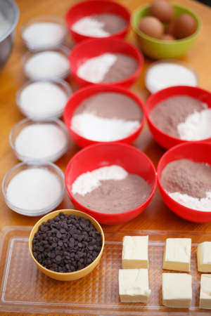 Cookery set of colorful bowls with chocolate drops, flour, cocoa powder, sugar, eggs and blocks of butter stand at light brown wooden table. Shallow dof. Focus on chocolate.の写真素材