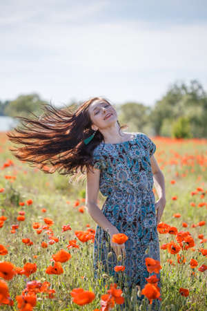 Young happy smiling woman with long brown hair in sundress with ethnic flower pattern stands on field of red poppies at the background of cloudy sky, trees and river with her hair flying in wind.の写真素材