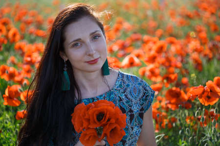 Portrait of young beautiful smiling woman in ornamented ethnic sundress and blue tassel earrings with long brown hair sitting on summer blooming flower field with bunch of poppies.の写真素材
