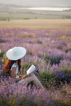 Young beautiful woman with long brown hair in dress and hat sits on lavender field with book in her hands under sunset light.の写真素材