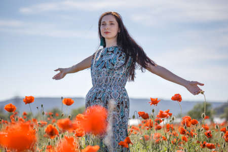 Young beautiful pensive woman with long brown flowing hair in sundress with ethnic flower pattern stands with open arms on field of red poppies at the background of cloudy sky.の写真素材