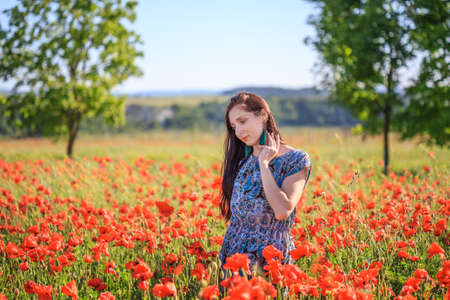 Beautiful thoughtful woman with long brown hair in sundress with ethnic flower pattern and tassel earrings stands on red poppy field and touches her hair.の写真素材