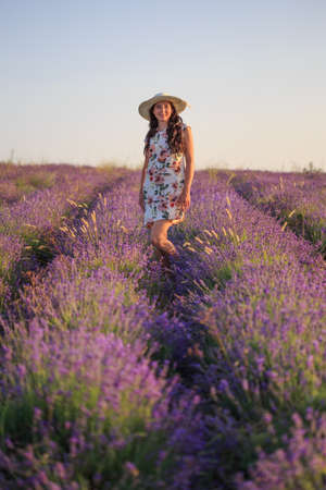 Young happy smiling romantic woman in light patterned sundress and hat stands on lavender field under sunset light.の写真素材