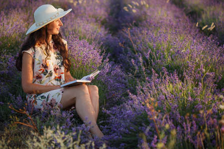 Beautiful romantic smiling woman with long brown hair in light patterned sundress and hat sits on lavender field with book in her hands and looks on sunset.の写真素材