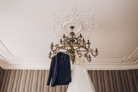 The groom's wedding suit hangs on the chandelier next to the bride's wedding dressの写真素材