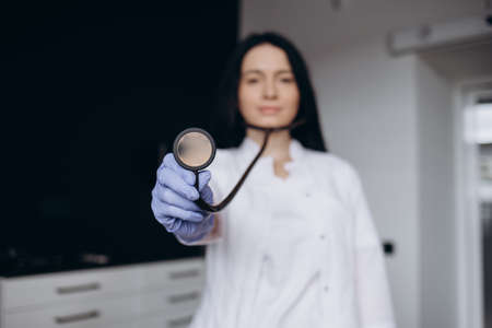 Close up smiling young Indian female doctor cardiologist holding stethoscope in hands, advertising regular professional cardiology checkup examination services for patients in modern clinic.の写真素材