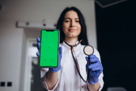 Close up smiling friendly young female doctor cardiologist holding cellphone with mockup screen and stethoscope, advertising mobile health tracking applications, telemedicine concept.の写真素材