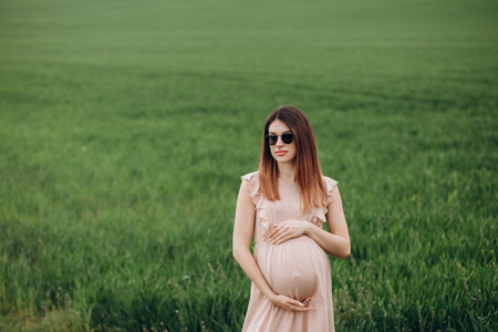 A beautiful young pregnant woman in the hat stands on a green field, leaning against a haystack. Sunny summer day. She is happy.の写真素材