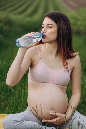 Pregnant woman drinking water after fitness. Thirsty after training expectant lady close-up, free space, sun light flareの写真素材