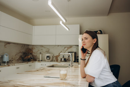 Young attractive smiling Latina woman talking on phone while cooking alone in kitchen. Housewife holds cellphone chatting distracted from healthy vegetarian food preparation. Chores, lifestyle concept.の写真素材