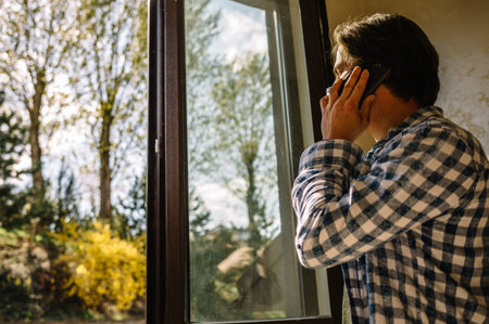 Young man wearing casual clothes talking on a mobile phone in the morning at a window with copy...