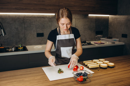 Beautiful young woman confectioner prepares sweets on a background of modern kitchenの写真素材