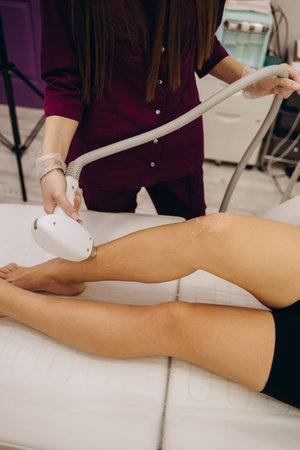 Doctors's hands in blue medical gloves holding a depilation machine and using it on the woman's legs. Focus is at the doctor's hands.Copy space.の写真素材