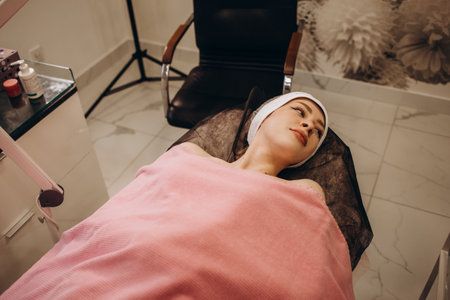 Curing skin problems. Cropped female cosmetologist looking at client's face through magnifying lamp examining her skin. Happy relaxed young woman getting professional facial treatment in spa salon.の写真素材