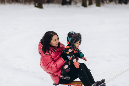 Happy mother with baby girl sitting on sledge and sledding down on snow from hill. Enjoying white winter day at the park. Spending time together in the weekend.の写真素材