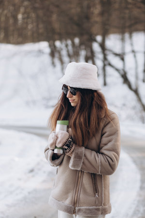 Smiling woman with dark hair in winter clothes with paper cup of coffee in hands against background of snowy winter forestの写真素材