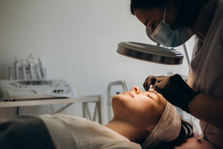 Middle-aged female cosmetician does eyebrows tweezing for elderly woman using special magnifying glassの写真素材