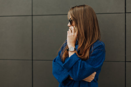 Businesswoman speaking over smart phone in the city. Beautiful in sunglasses and long blue coat holding white little bag at her hand.の写真素材