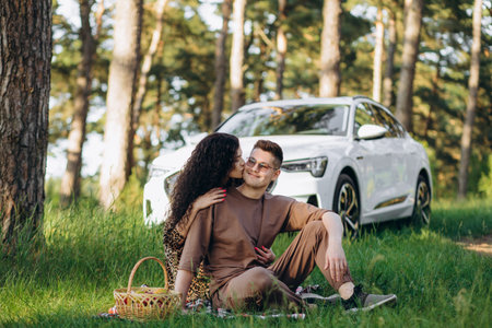 Young modern couple enjoying a picnic in nature, sitting near the parked white electric carの写真素材