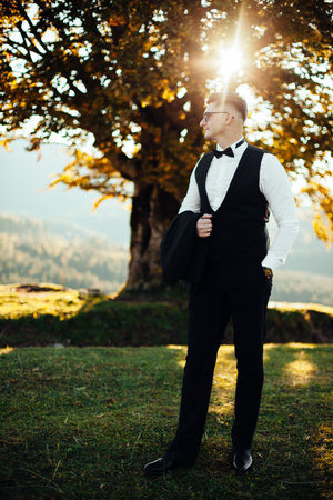 Groom in a stylish suit walking in the field at sunset. Happy smiling Caucasian man in white shirt, black vest and beige pants standing in the field outdoorsの写真素材