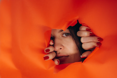 surprise. Portrait of a young caucasian woman looking through the hole in orange paper. Distrustful look. Women's curiosity and gossip. A jealous wife. copyspace.の写真素材