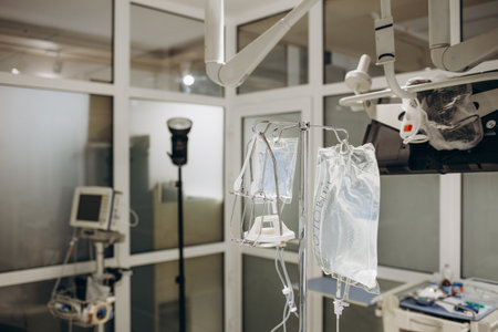 IV infusion bags and bottles hanging on poles during a real surgery going on in the background. Patient, illness, treatment, anesthesia, hospital, medicine and healthcare symbolism and concept.の写真素材