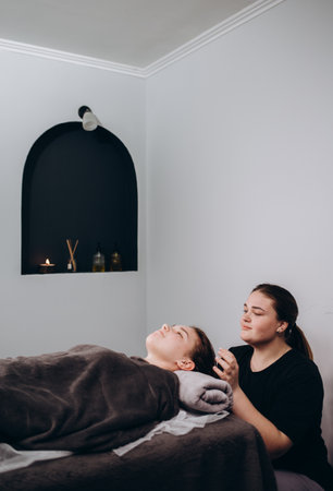young beautiful spa customer laying down closed eyes covered with sliced organic cucumber on massaging bed while unrecognizable professional female masseuse therapist massage head and face.の写真素材