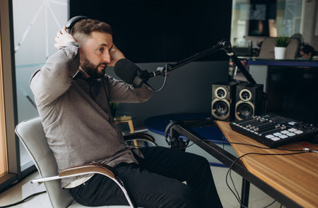 Handsome young latin man working as radio host at radio station sitting in front of microphone holding clipboard and using mobile phoneの写真素材