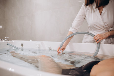 Young woman during hydromassage in beauty salon. Concept of body care and spa. Masseuse massages the girl in the bath with a jet of waterの写真素材