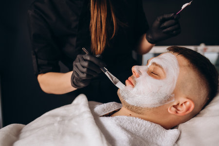 Man getting facial nourishing mask by beautician at spa salon, closeup. Apply face mask, spa beauty treatment and skincare.の写真素材