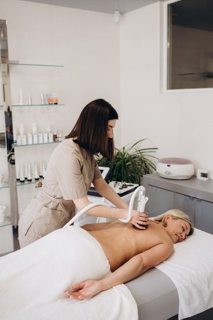 A therapist massages a woman's back with a massage percussion device in her home. The therapist's hand holds a therapeutic vibrating massager. Physical therapy and muscle recovery massageの写真素材